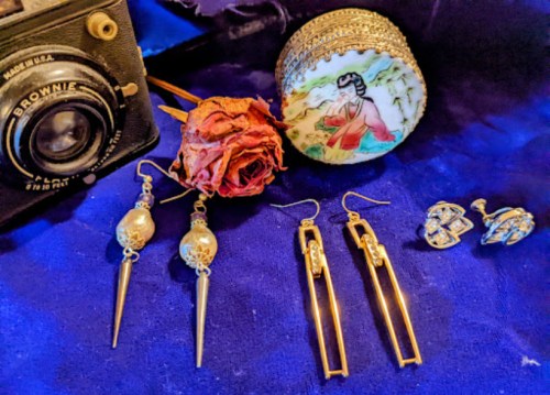 A vintage camera, decorative box, dried rose, and various earrings on a blue fabric background.