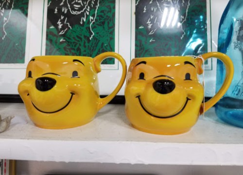 Two yellow mugs shaped like smiling cartoon bears, displayed on a shelf with framed artwork in the background.