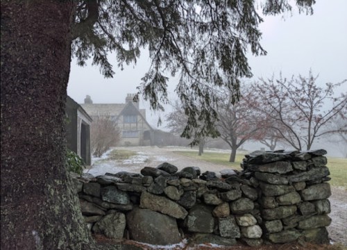 A foggy landscape featuring a stone wall, trees, and a distant house partially obscured by mist.