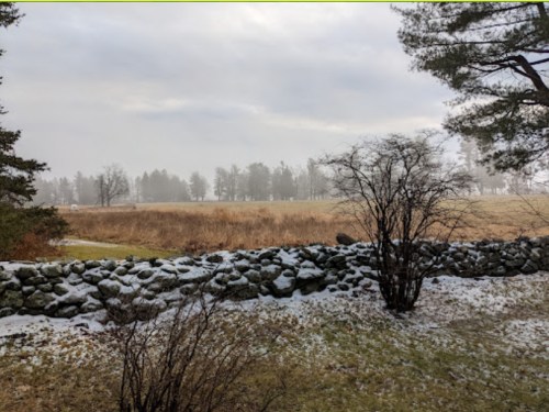 A misty landscape featuring a stone wall, barren trees, and a field with patches of snow under a cloudy sky.