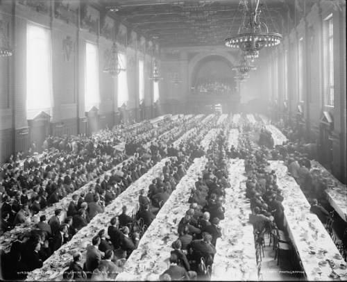 A large banquet hall filled with rows of tables set for a formal dinner, crowded with elegantly dressed guests.