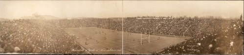 Historic panoramic view of a large crowd at a rugby match, with players on the field and mountains in the background.
