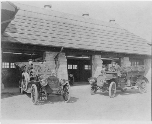 Two vintage cars parked in front of a stone garage, with two men seated in the vehicles.