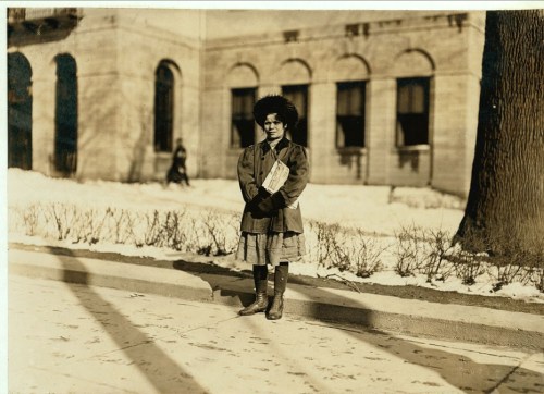 A young girl stands in winter attire on a snowy street, with a building and another person in the background.