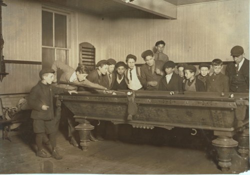 A group of boys gathered around a billiard table in a vintage setting, focused on the game.