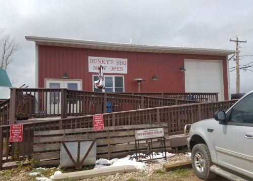 A red building with a sign reading "Bunky's BBQ Now Open," featuring a wooden deck and parked vehicle in front.