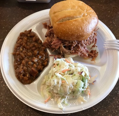 A plate with a pulled pork sandwich, baked beans, and coleslaw on a disposable tray.