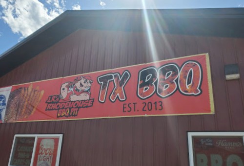 Sign for JR's Rhodehouse BBQ Pit, featuring "TX BBQ" and established in 2013, on a red building with a blue sky.