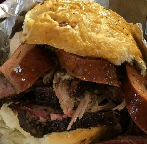 A close-up of a hearty sandwich filled with smoked meats, including brisket and sausage, on a toasted bun.