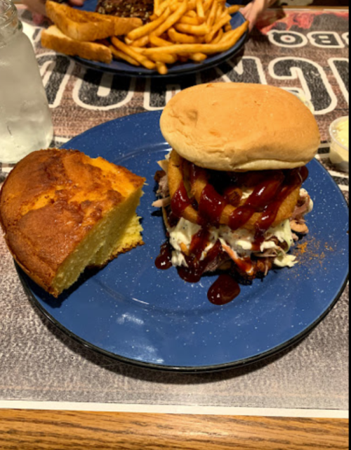 A plate featuring a stacked BBQ sandwich with sauce and onion rings, alongside a slice of cornbread.