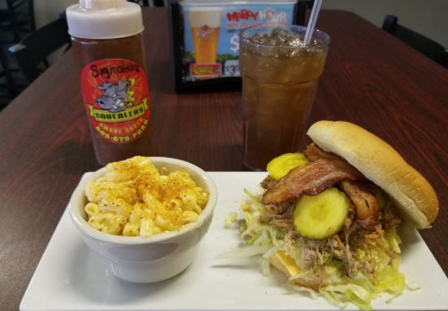 A plate with a sandwich topped with bacon, pickles, and shredded lettuce, alongside macaroni and cheese and a drink.