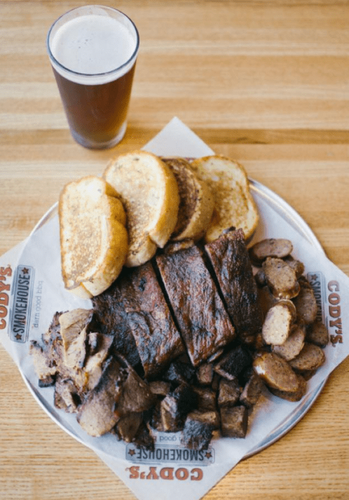 A platter of smoked meats including ribs, sausage, and brisket, served with slices of bread and a pint of beer.