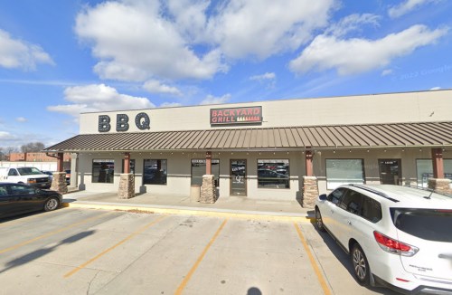 Exterior of a restaurant building with a sign reading "BBQ" and "Backyard Grill," featuring a parking lot and blue sky.