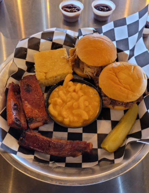 A metal tray with two pulled pork sandwiches, macaroni and cheese, cornbread, ribs, a pickle, and barbecue sauce.
