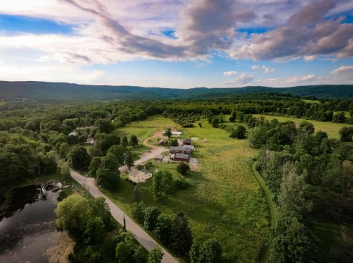 Aerial view of a lush green landscape with a farm, winding road, and a pond surrounded by trees under a cloudy sky.
