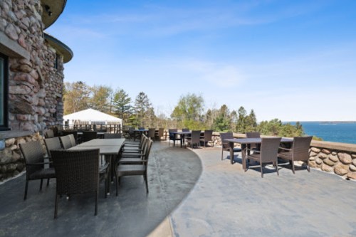 Outdoor dining area with stone walls, tables, and chairs overlooking a scenic view of the water and trees.