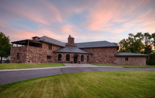 A large stone house with a sloped roof, surrounded by green grass and trees, under a colorful sunset sky.