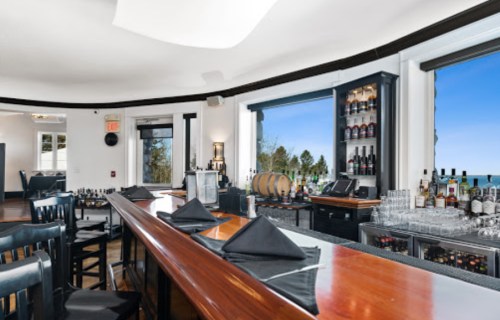 A stylish bar with a polished wooden counter, black napkins, and large windows showcasing a scenic view.