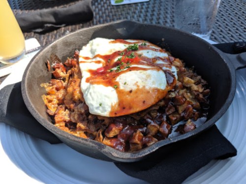 A cast iron skillet filled with crispy potatoes, topped with a fried egg and drizzled with sauce, served on a white plate.