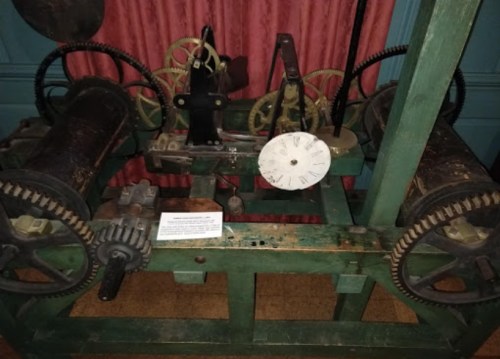 A vintage mechanical device with gears and dials, displayed on a wooden base against a red curtain backdrop.