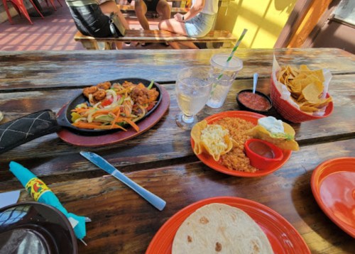 A table with a sizzling dish of shrimp and vegetables, rice, tortillas, chips, salsa, and drinks in a vibrant setting.