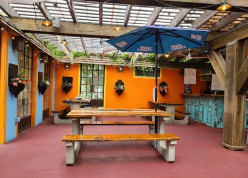 Outdoor seating area with wooden tables, colorful walls, and an umbrella, surrounded by hanging plants and rustic decor.