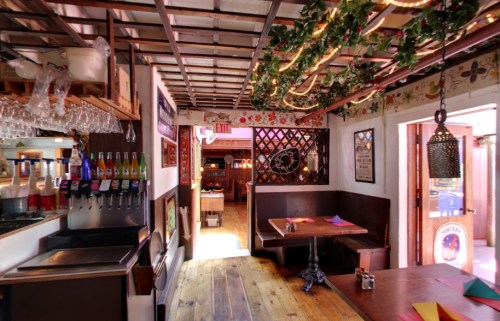 Cozy restaurant interior with wooden floors, colorful decor, and a bar area featuring drinks and glassware.