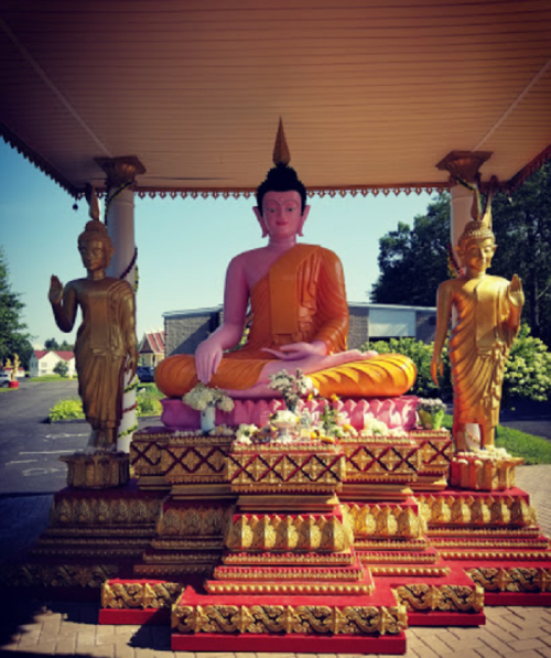 A pink Buddha statue seated on a pedestal, flanked by two golden figures, under a covered structure in a garden setting.