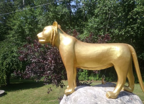 A golden lion statue stands on a rock, surrounded by greenery and trees in the background.