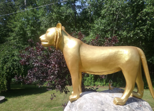 A golden lion statue stands on a rock, surrounded by greenery and trees in the background.