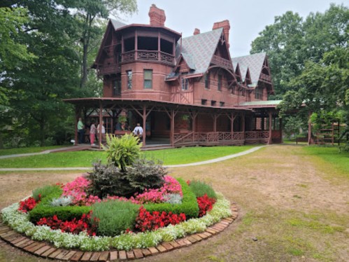 A large, historic red mansion with intricate architecture, surrounded by a colorful flower garden and green trees.