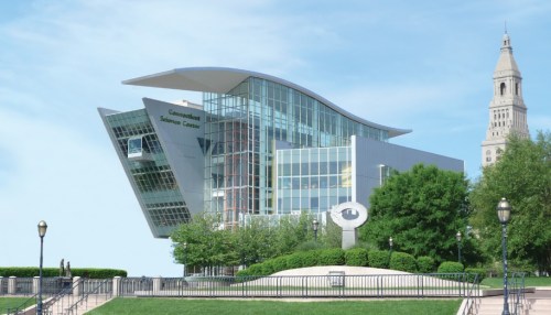 Modern building with a unique design, surrounded by greenery and a clock tower in the background under a clear blue sky.
