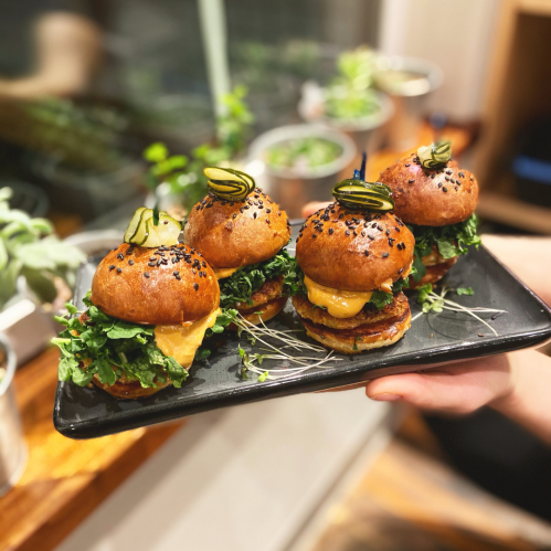 A platter of four mini burgers with lettuce, cheese, and pickles, garnished with sesame seeds and microgreens.