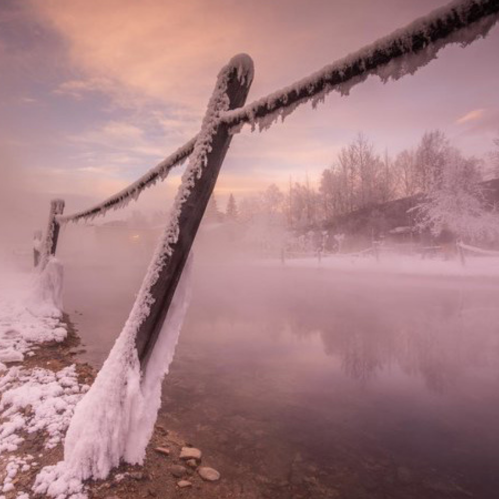 A frosty wooden fence post by a misty river, surrounded by snow-covered trees and a soft, colorful sky at dawn.