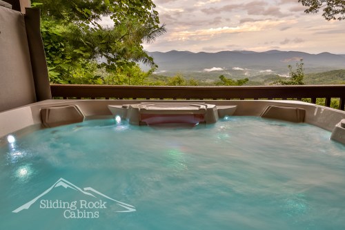 A hot tub with glowing lights overlooks a misty mountain landscape at sunset.