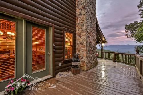 A cabin exterior with stone and wood features, a deck, and mountain views at sunset. Flowers adorn the entrance.