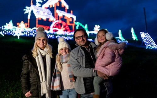 A family of four stands together in front of colorful holiday lights on a dark evening.