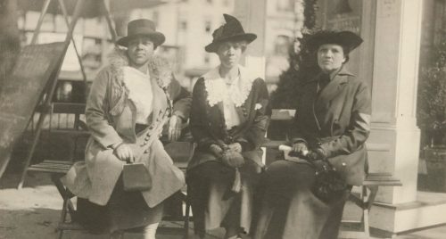 Three women in early 20th-century attire sit together on a bench, each wearing hats and holding umbrellas.