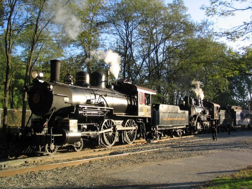 Two vintage steam locomotives with billowing smoke, surrounded by trees and a gravel path.