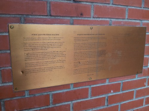 A bronze plaque mounted on a brick wall, featuring text in two columns.