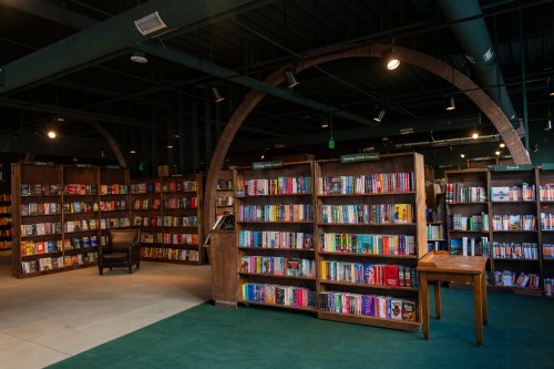 A cozy bookstore with wooden shelves filled with books, green carpet, and a chair in a well-lit space.