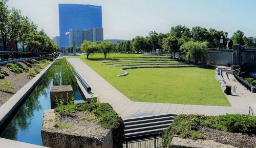A landscaped park with green grass, water features, and modern buildings in the background under a clear blue sky.