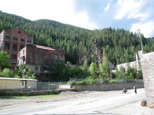 Abandoned industrial buildings surrounded by trees and mountains, with a gravel road in the foreground.
