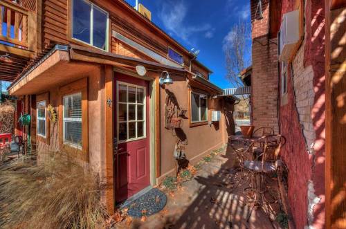 A cozy outdoor area with a red door, brown walls, and a small patio featuring two chairs and a table under a blue sky.