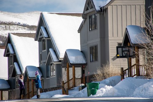 Two people stand on a snowy street near houses with sloped roofs, surrounded by winter scenery.