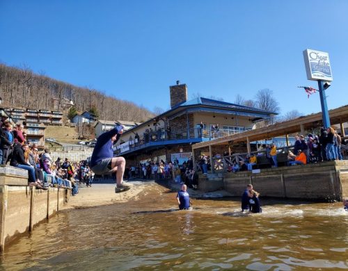 A person jumps into a lake while others watch from the shore, enjoying a sunny day at a marina event.