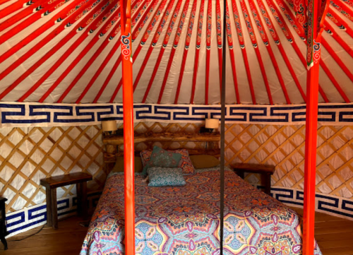 Interior of a yurt featuring a colorful patterned bedspread, wooden furniture, and a vibrant red ceiling.