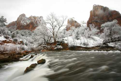 A snowy landscape with a flowing river, surrounded by rocky cliffs and bare trees in a serene, winter setting.