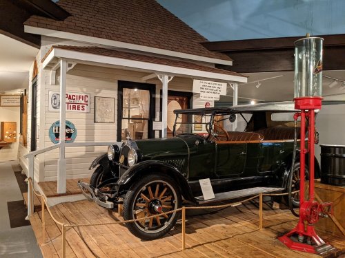 A vintage green car displayed in a museum, next to a historic gas pump and a replica of a small building.