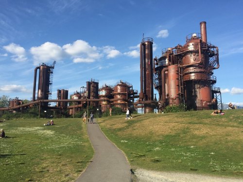 Industrial structures with rusted pipes and towers, set against a blue sky, surrounded by green grass and people walking.